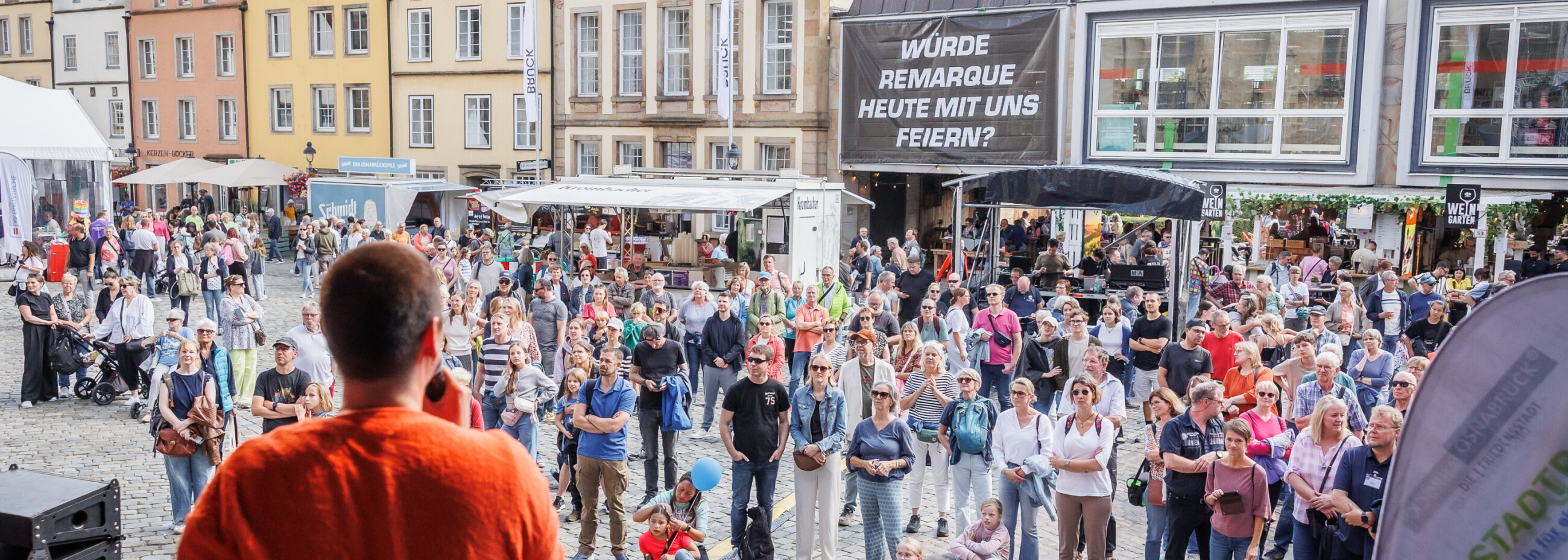 Blick von der Bühne auf den Moderator in orangem Shirt, der vor großem Publikum auf dem Osnabrücker Marktplatz beim Charity Stadtradeln 2025 spricht.