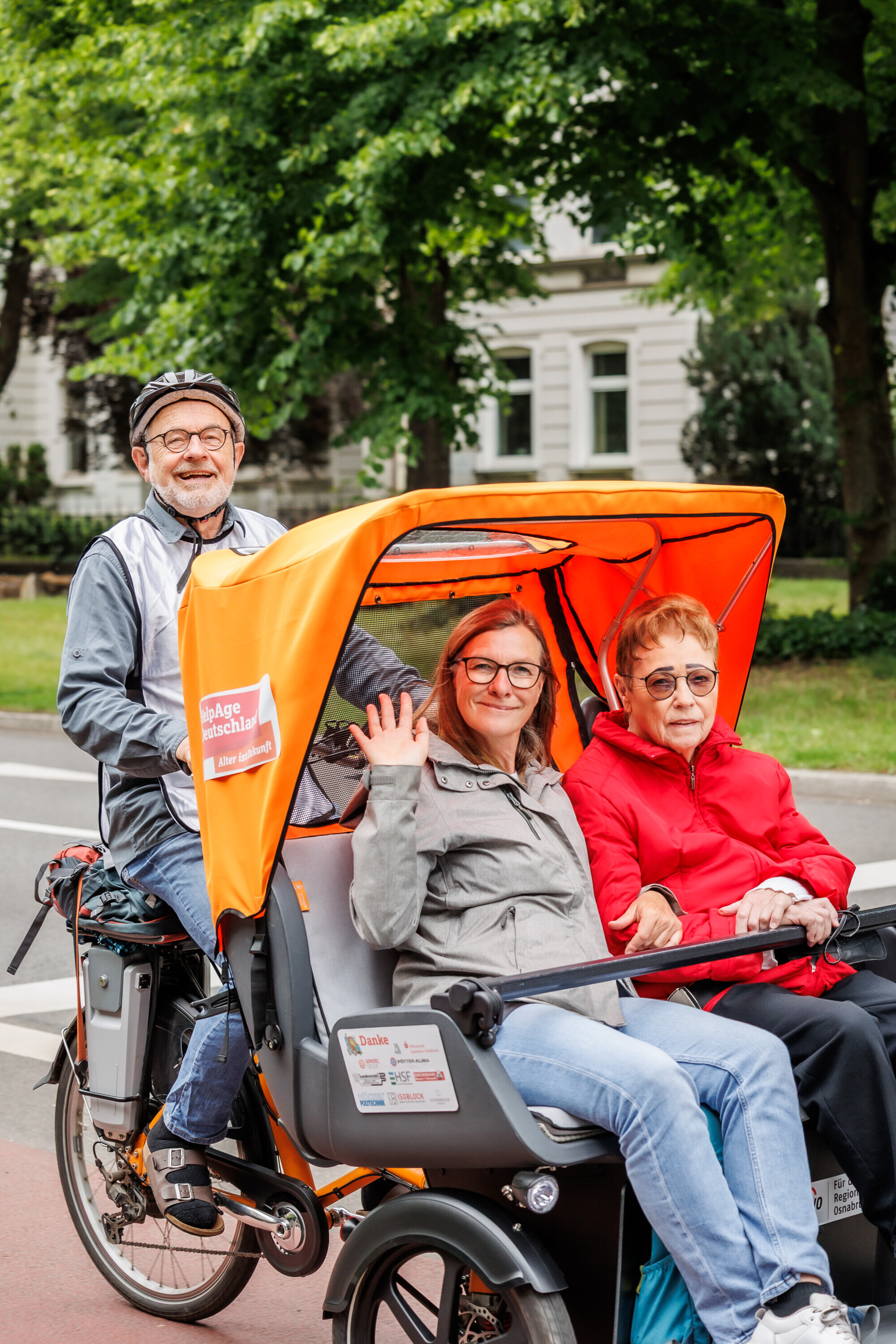 Ein älterer Mann fährt eine orangefarbene Rikscha mit zwei Mitfahrerinnen, eine davon winkt freundlich in die Kamera.
