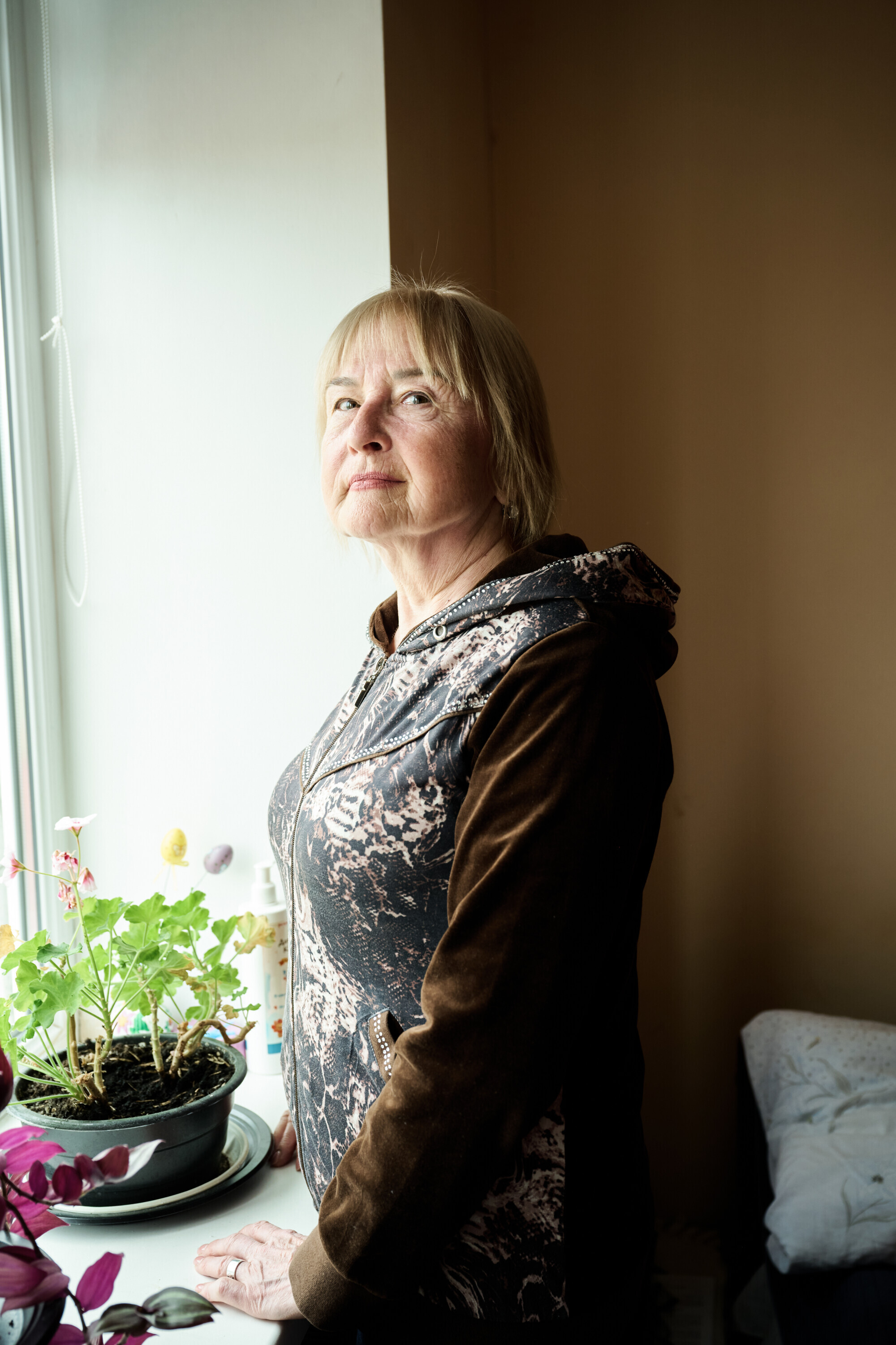 Ukrainische Seniorin steht nachdenklich an einem Fenster mit Blumentopf – Hoffnung und Würde im Alltag des Krieges.