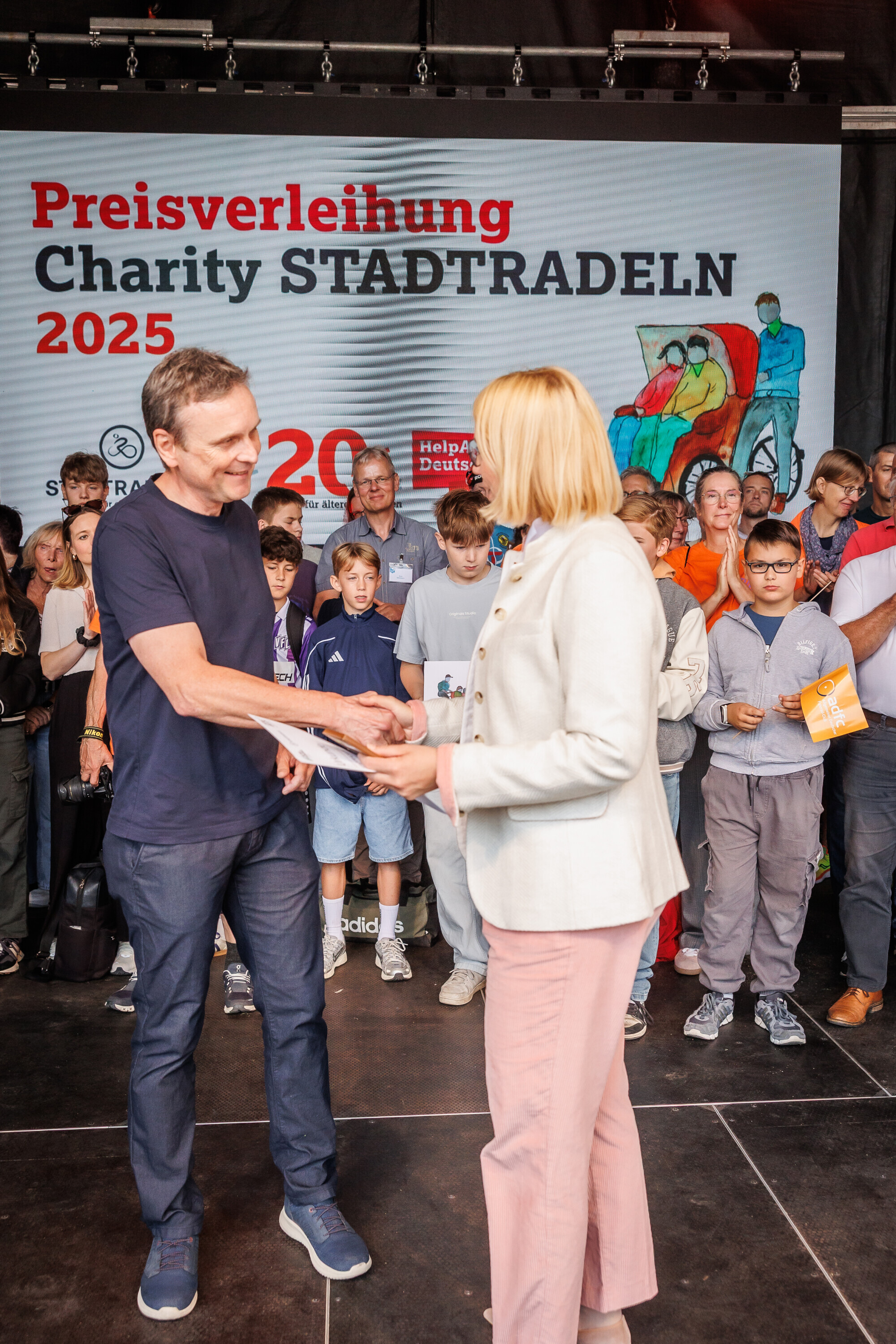 Sven Frommhold nimmt bei der Preisverleihung des Charity Stadtradelns 2025 eine Auszeichnung entgegen und schüttelt die Hand einer Moderatorin.