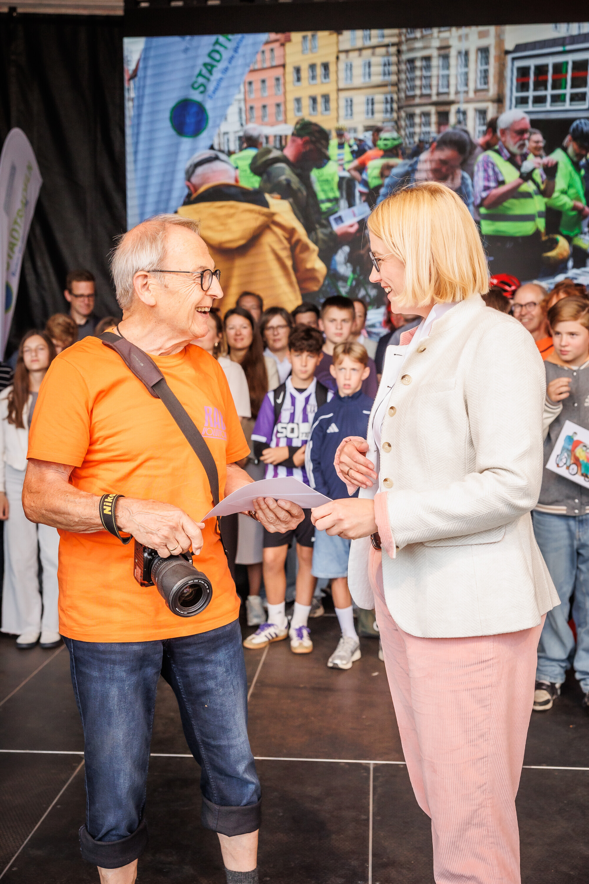 Rainer Schiemann lacht bei der Übergabe seiner Urkunde beim Charity Stadtradeln 2025 und hält eine Kamera in der Hand.