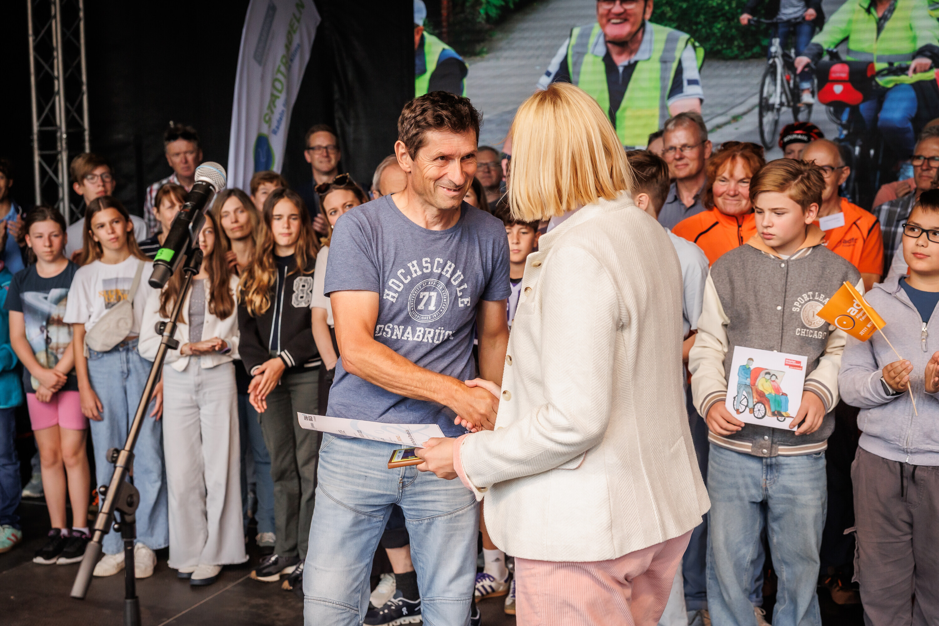 Ein Teilnehmer im T-Shirt der Hochschule Osnabrück nimmt eine Urkunde beim Charity Stadtradeln 2025 entgegen und schüttelt die Hand der Moderatorin.