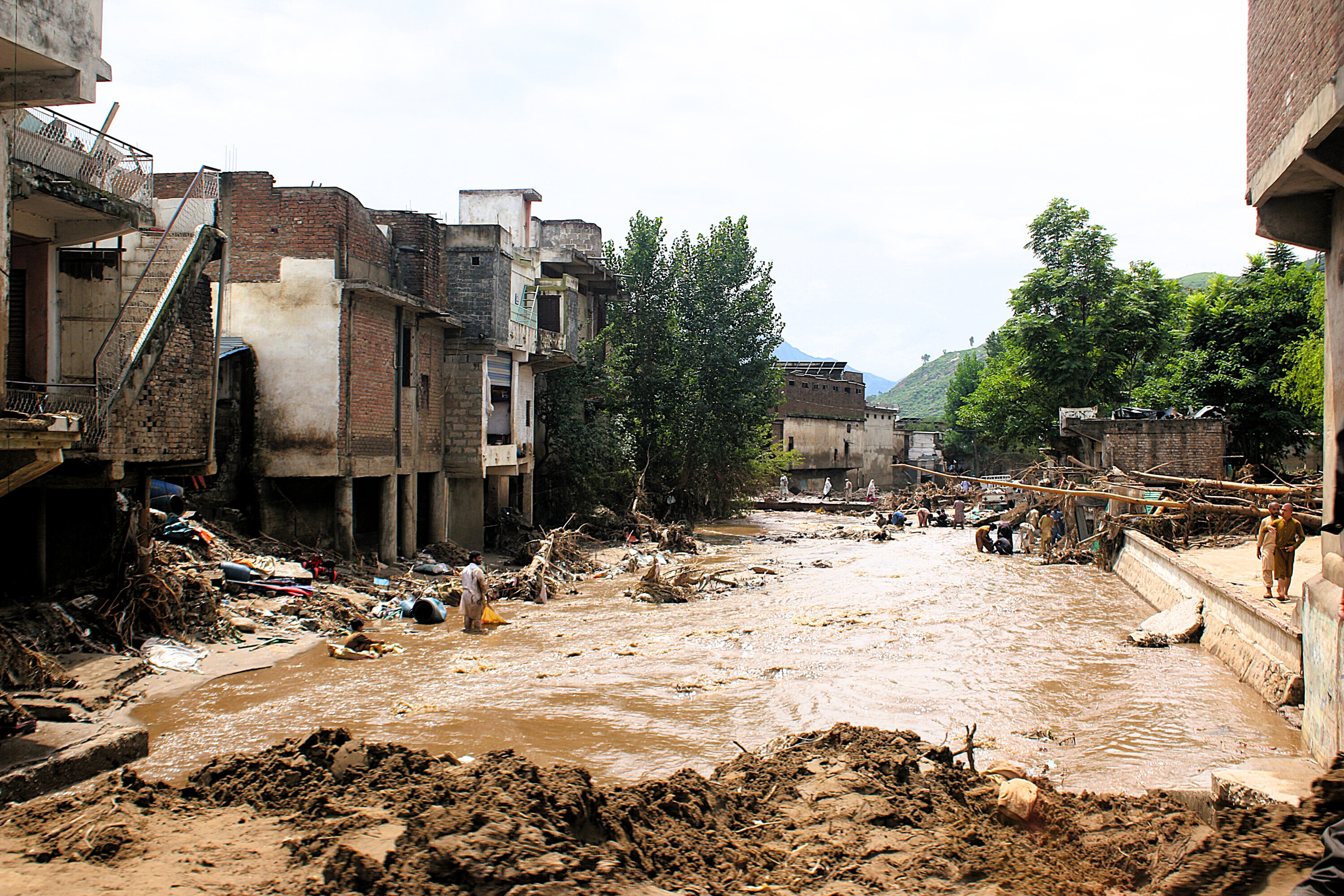 Überflutete Straße in Pakistan, umgeben von beschädigten Häusern, Trümmern und Menschen, die sich durch das Hochwasser bewegen.
