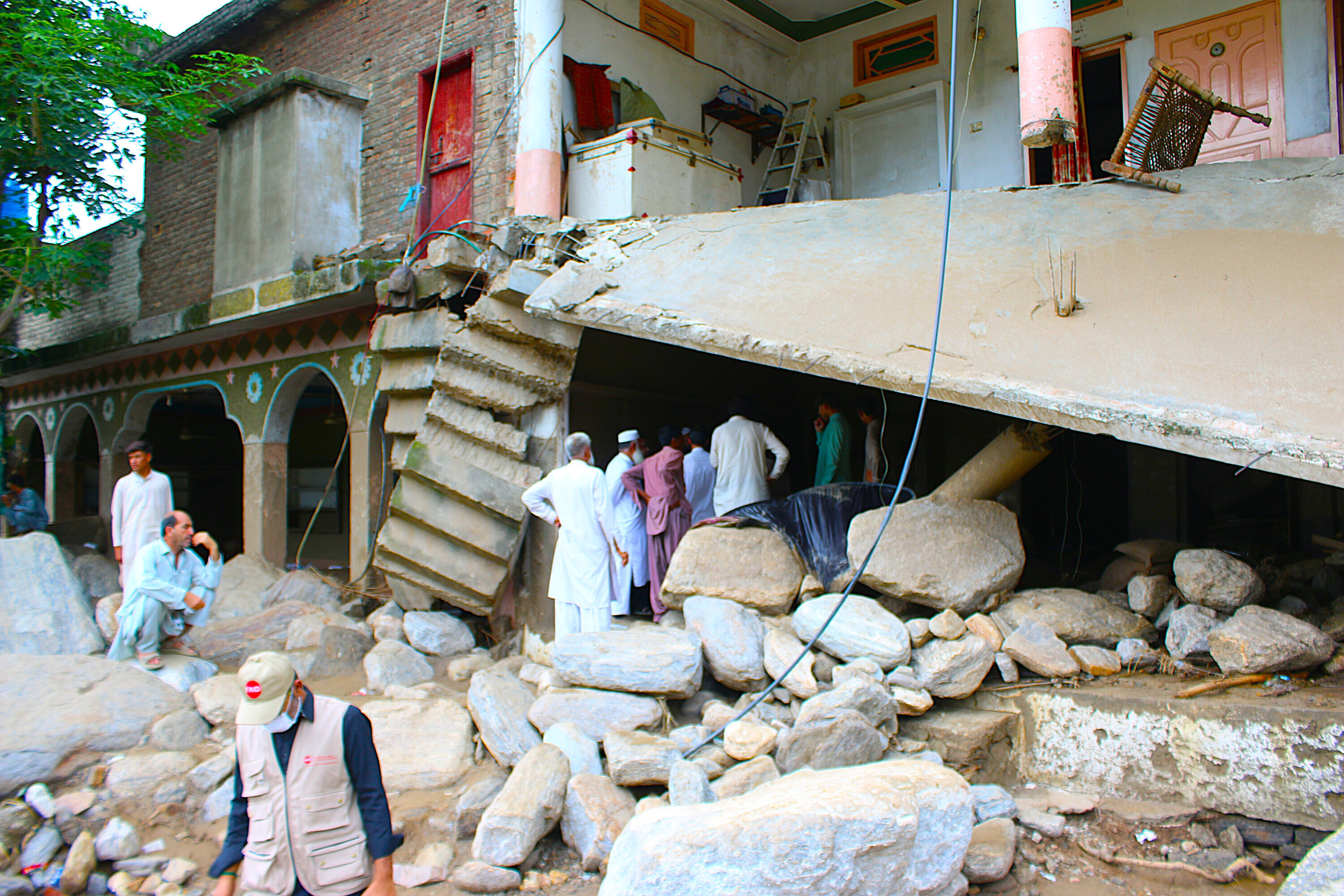 Eingestürztes Wohnhaus in Pakistan nach Sturzfluten, Felsen und Trümmer im Vordergrund, während Menschen die Schäden begutachten.