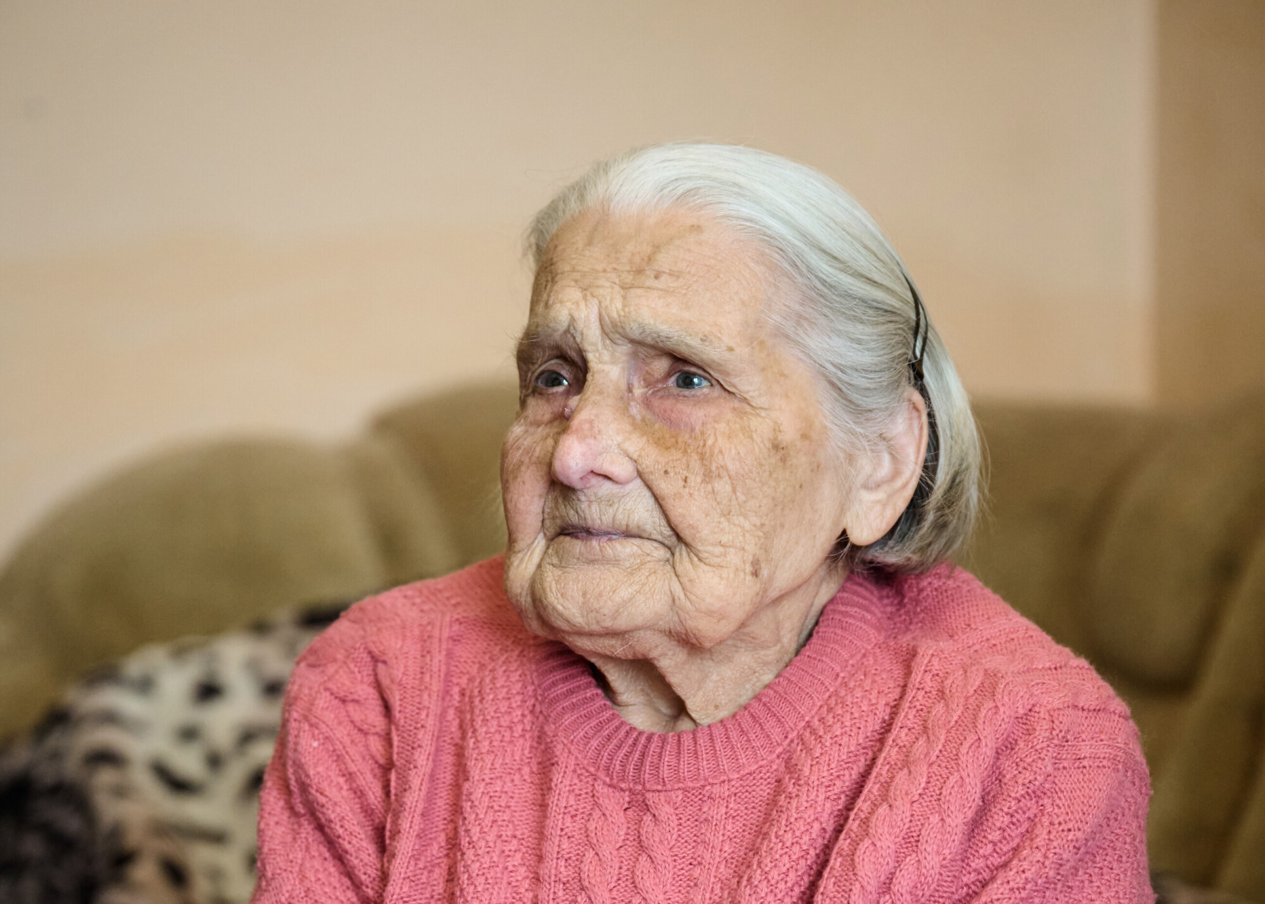97-jährige Frau mit rosa Strickpullover sitzt in ihrer Wohnung – gezeichnet vom Leben, aber voller Ausdruckskraft.