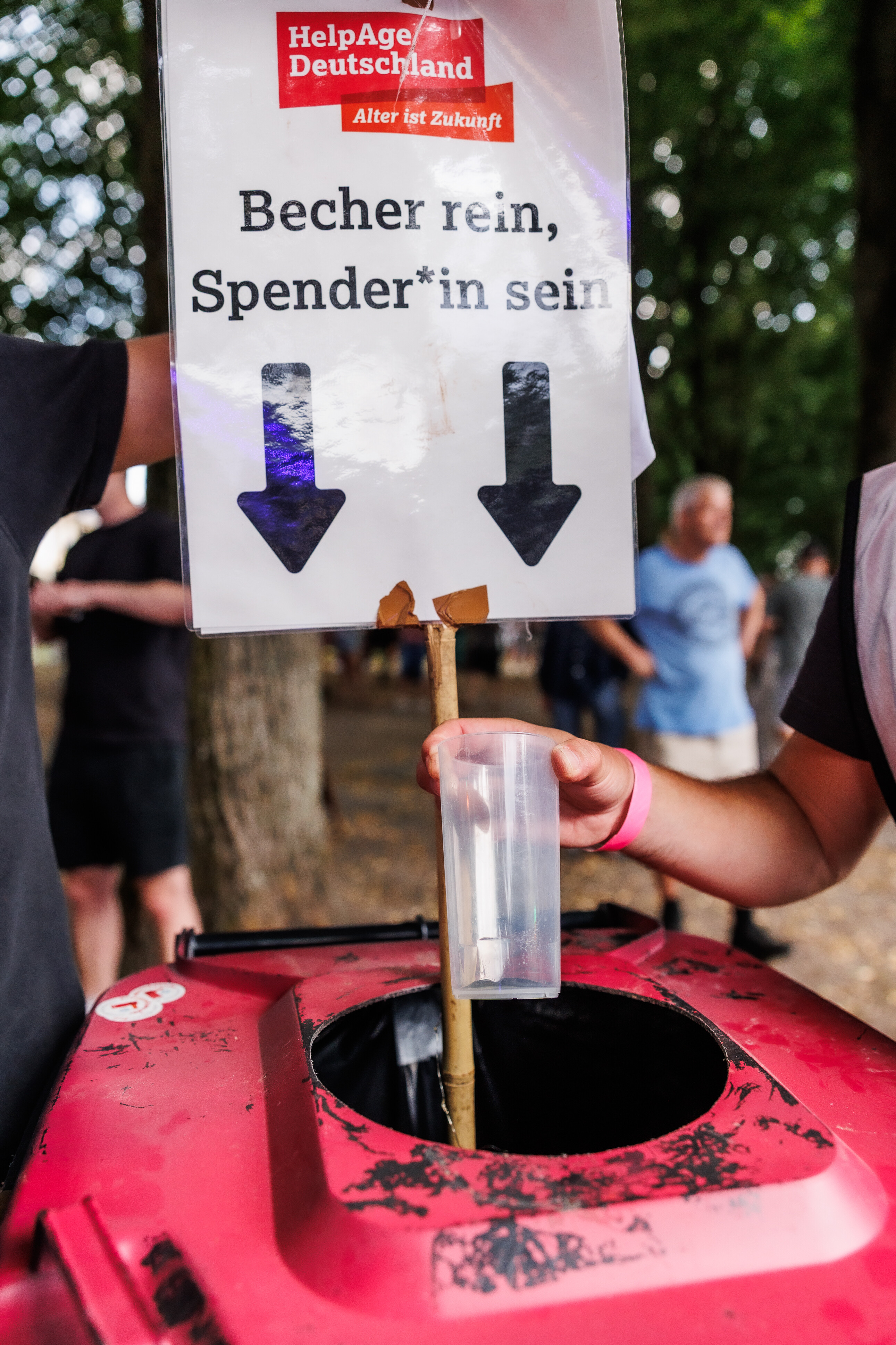 Ein Festivalbesucher wirft einen Pfandbecher in eine rote Tonne mit dem Schild „Becher rein, Spenderin sein“ von HelpAge Deutschland.