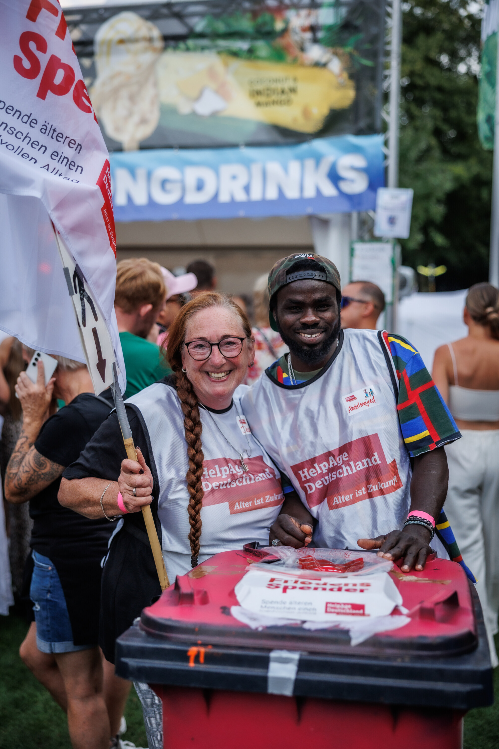 Zwei freiwillige Helferinnen von HelpAge Deutschland lächeln in die Kamera und stehen hinter einer roten Spendentonne beim Festival.