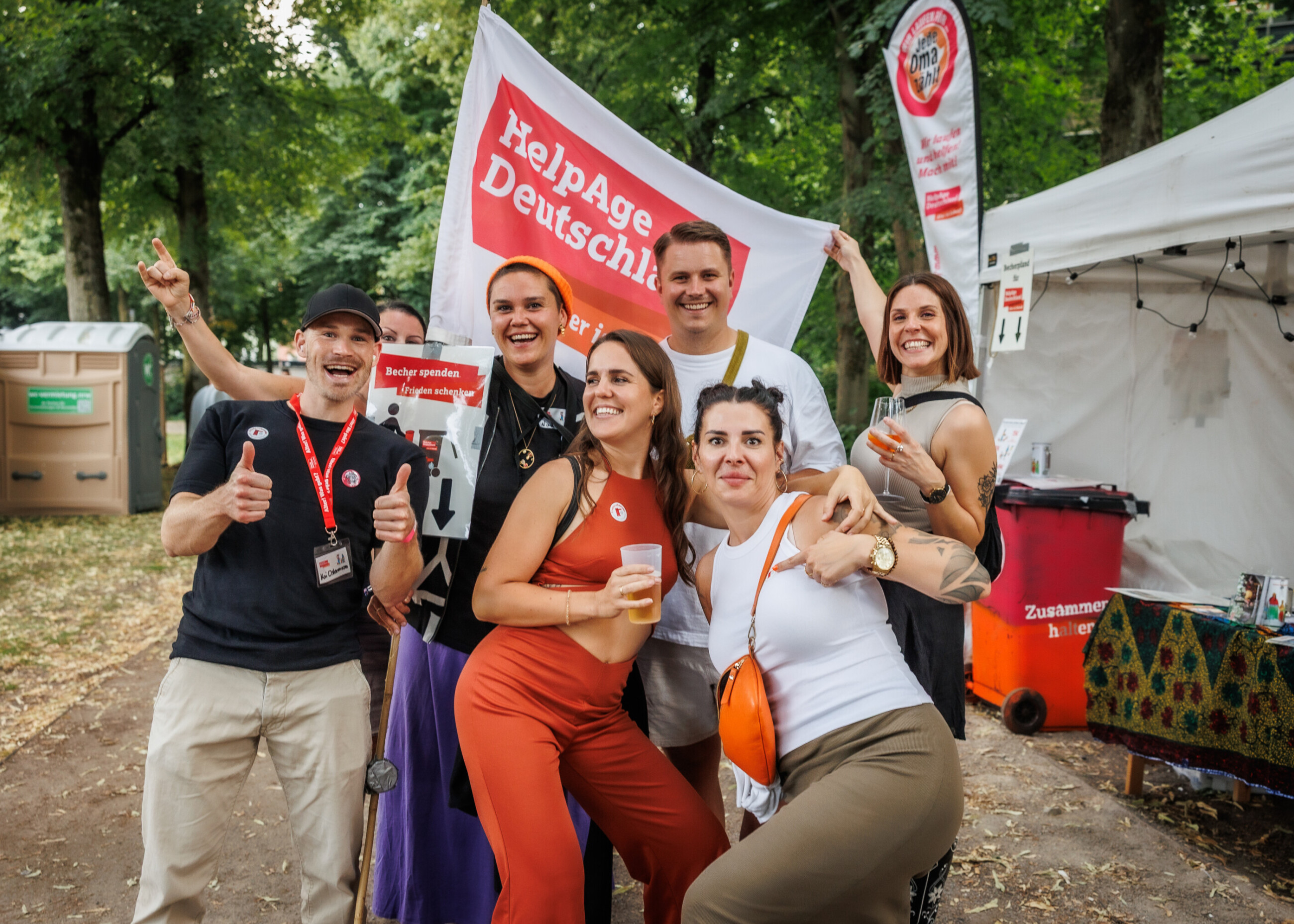Eine Gruppe junger Festivalbesucherinnen posiert fröhlich mit Fahne und Schild von HelpAge Deutschland neben einer roten Spendentonne.