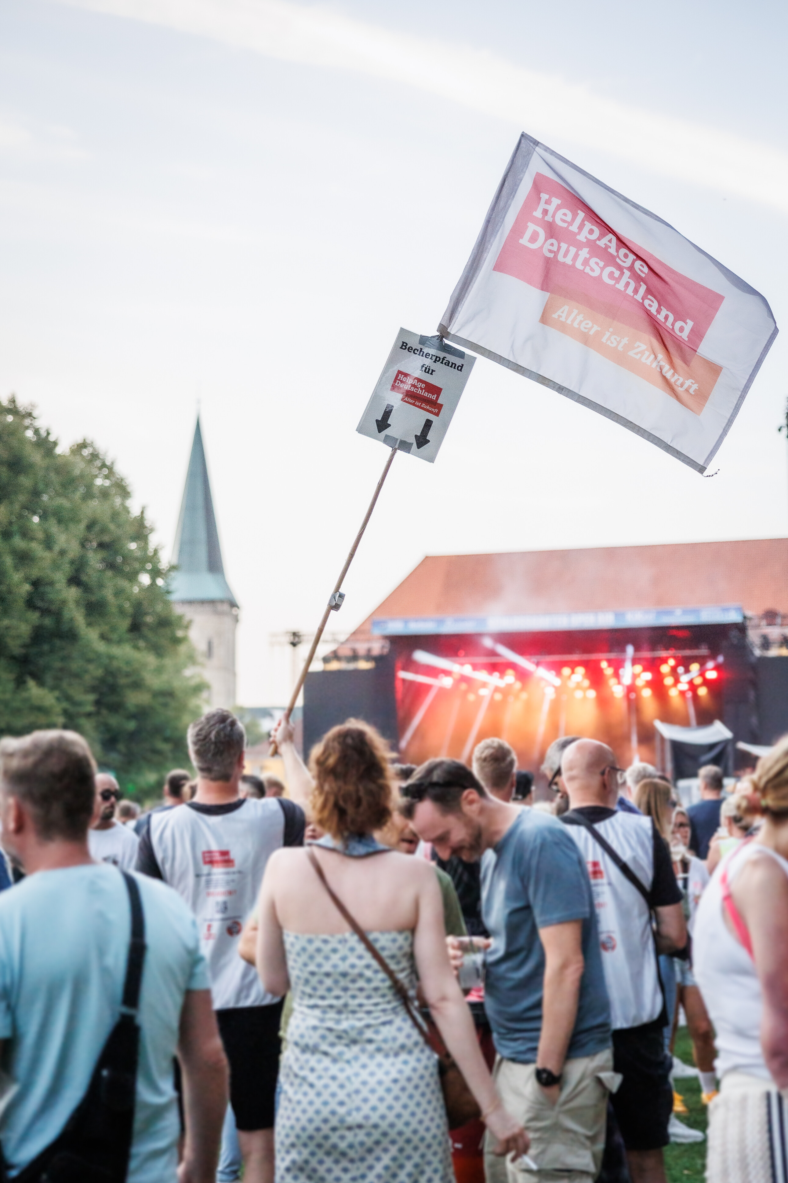 Festivalbesucherinnen stehen vor einer Bühne, während eine große HelpAge Deutschland-Fahne mit Becherspenden-Hinweis in die Höhe gehalten wird.