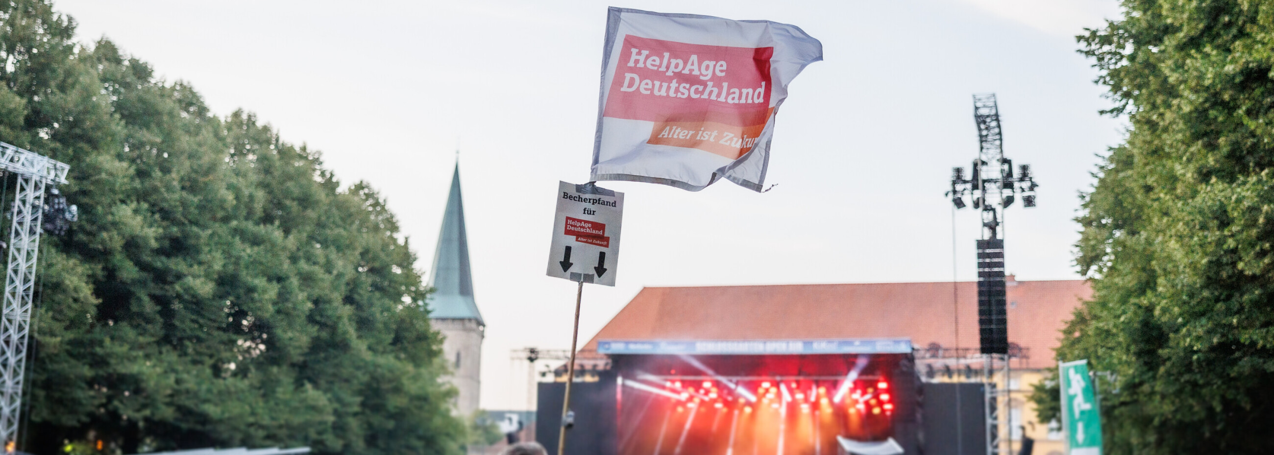 Blick über die Menschenmenge vor der Festivalbühne, im Vordergrund eine HelpAge Deutschland-Fahne mit Becherspende-Aufschrift.